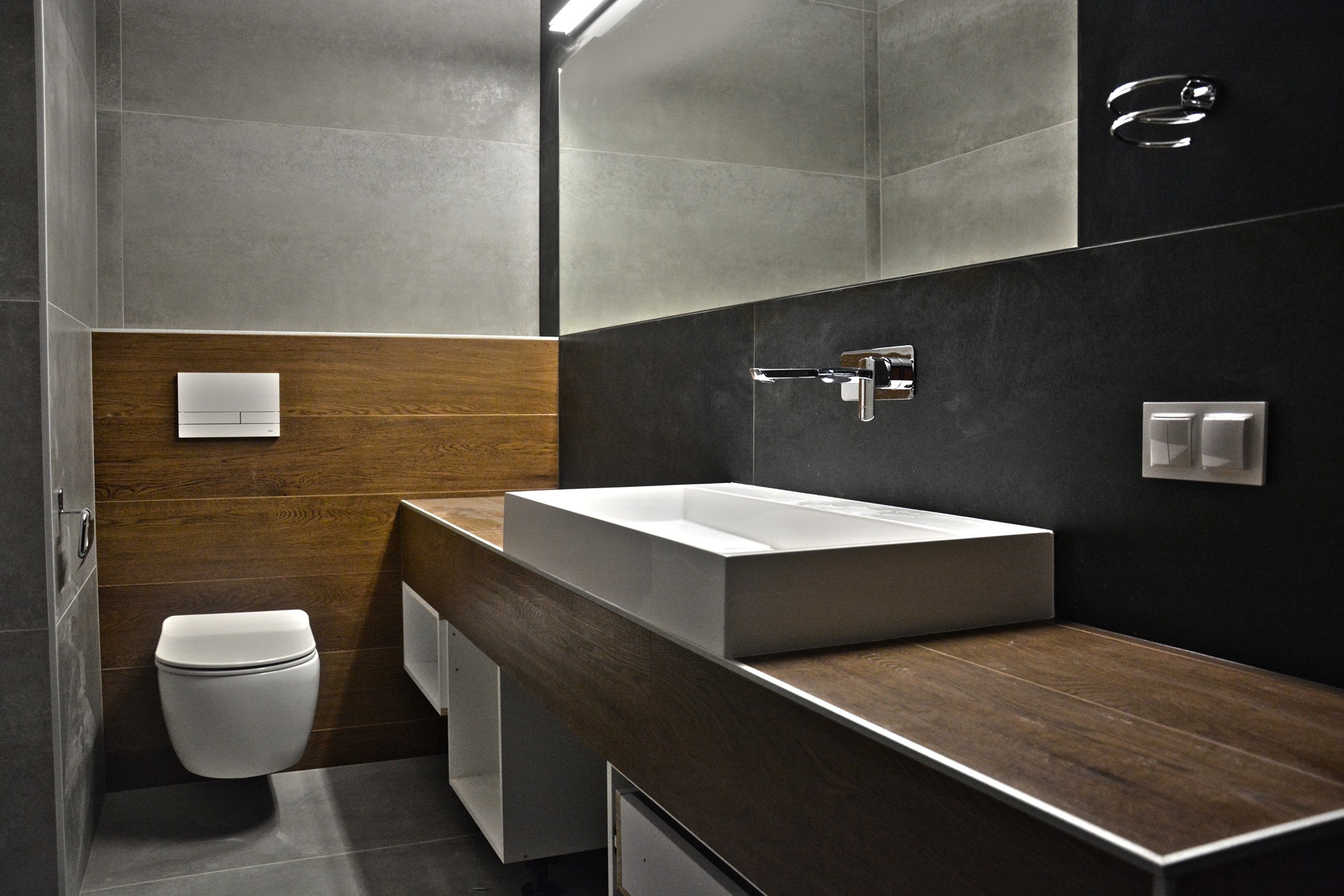 Nowoczesna łazienka z drewnianymi akcentami: prostokątna umywalka nablatowa, szare płytki i podwieszana toaleta. Minimalistyczny design z oświetleniem LED.