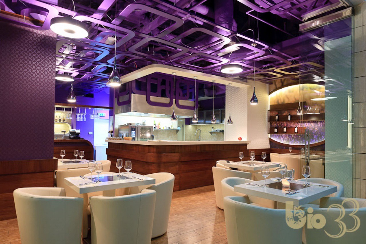 Nowoczesne wnętrze restauracji z fioletowym oświetleniem, białymi krzesłami i drewnianymi elementami. Widoczne nakryte stoły i bar.