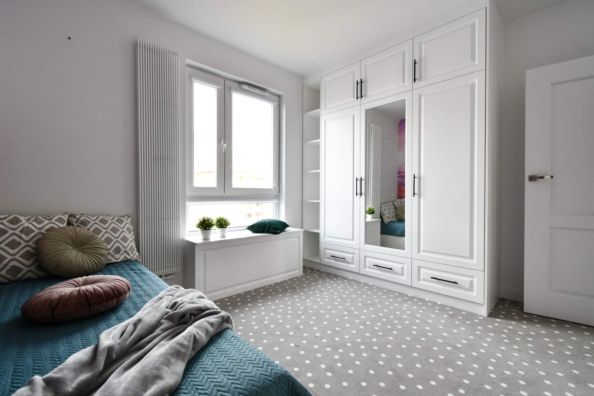 Jasna sypialnia z zabudową szafy z lustrem, łóżkiem z dekoracyjnymi poduszkami i dywanem w kropki. Minimalistyczny design z akcentami kolorystycznymi.