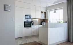 Jasna kuchnia z zabudową na wymiar: kremowe szafki, sprzęt AGD w zabudowie, marmurowa podłoga i minimalistyczny zegar na ścianie. Nowoczesny design.