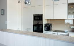 Jasna kuchnia z zabudową na wymiar: białe szafki, sprzęt AGD (piekarnik, mikrofalówka, ekspres do kawy) i blat z subtelnym wzorem. Minimalistyczny design.