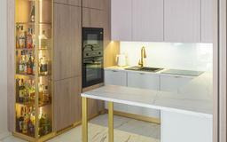 Nowoczesna kuchnia z barkiem: beżowe fronty, złote akcenty, oświetlona witryna z alkoholem, marmurowy blat i czarny piekarnik w zabudowie.