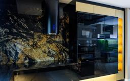 Nowoczesna kuchnia: czarne, lakierowane fronty szafek, blat z granitu o wyraźnym użyleniu, podświetlenie LED i czarny okap cylindryczny.