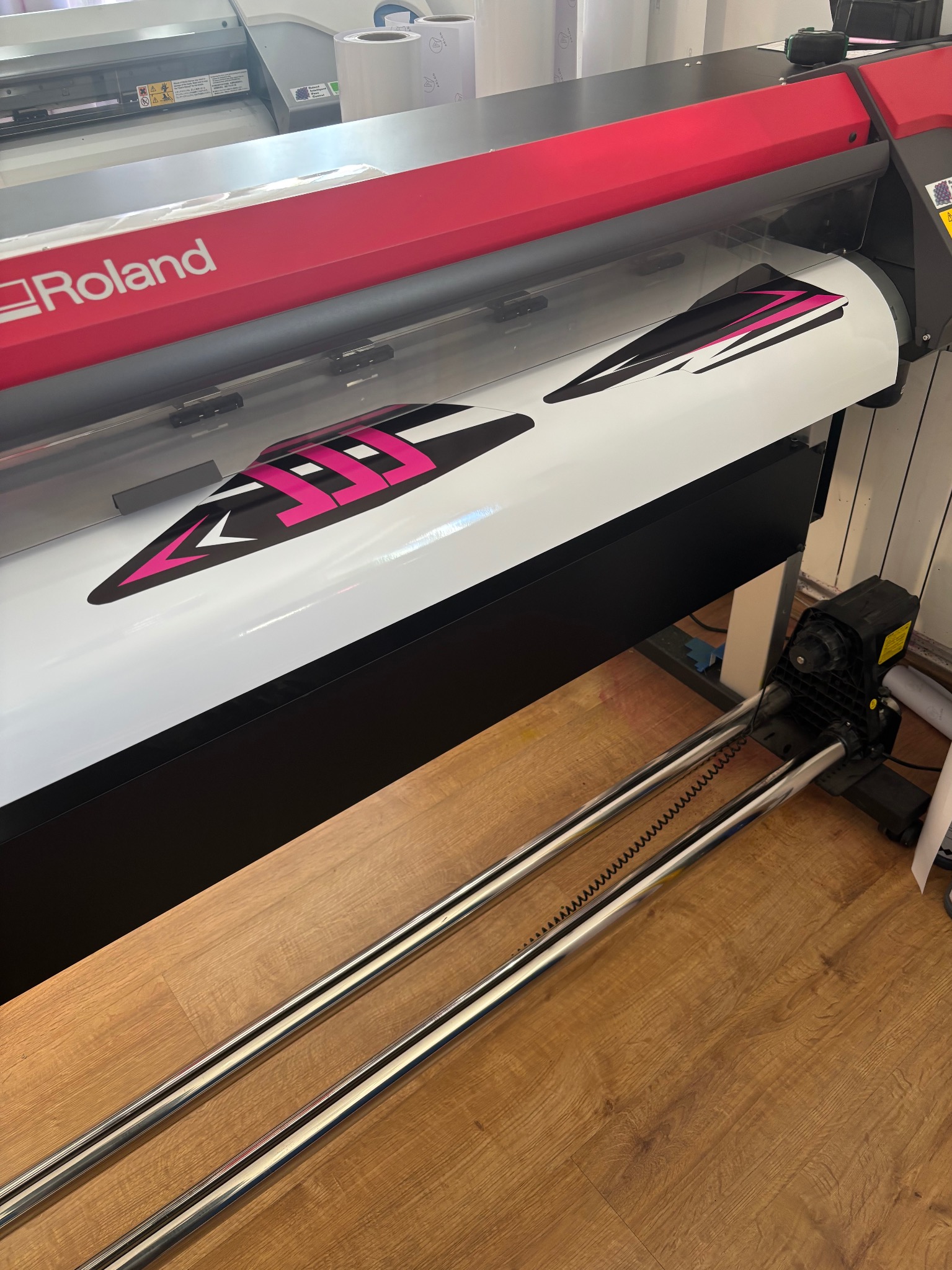 Drukarka wielkoformatowa Roland drukuje naklejki z czarno-różowym wzorem na białym tle. Widoczne rolki z folią i elementy mechaniczne urządzenia.