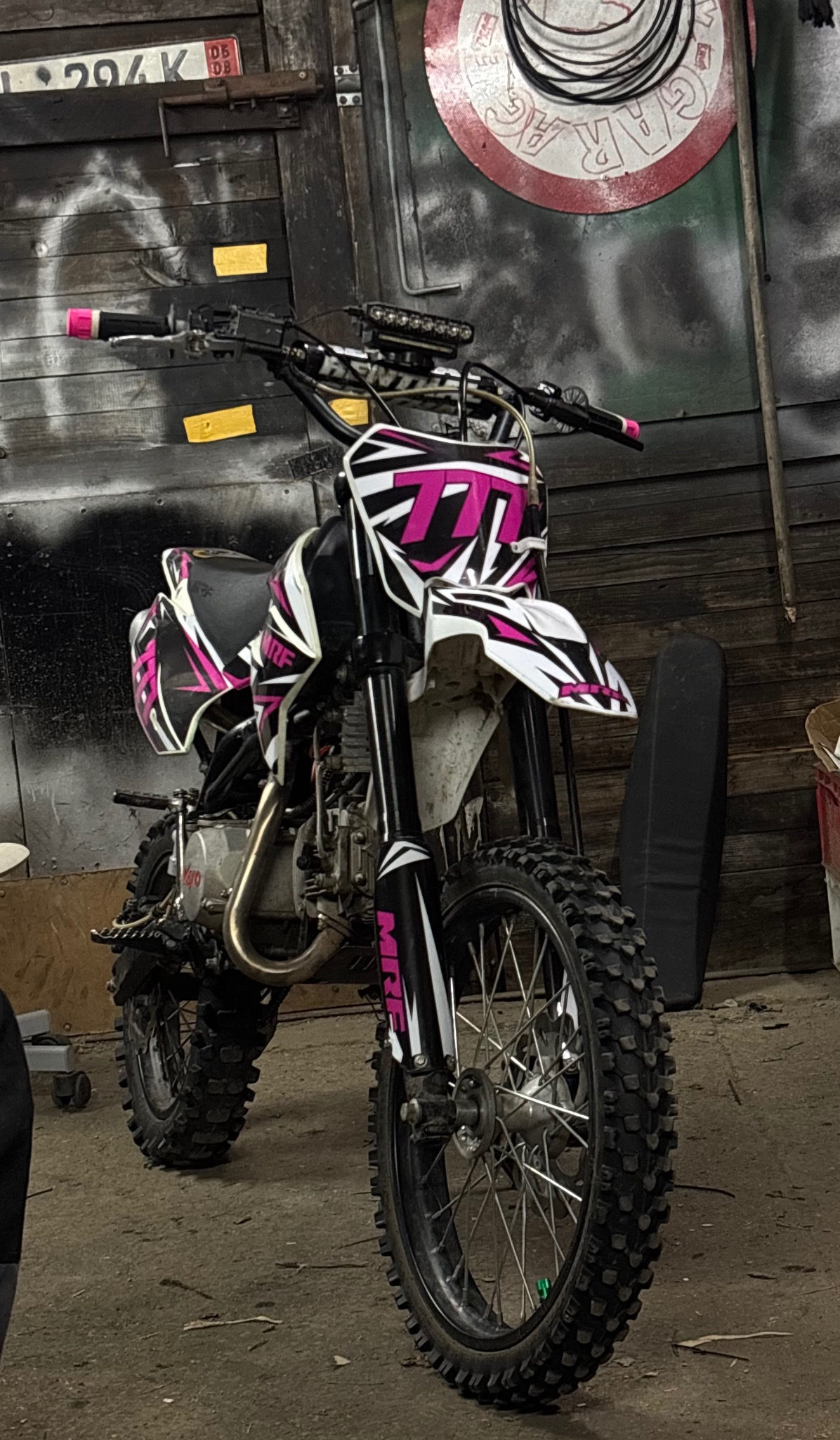 Dirt bike MRF z różowymi akcentami i grafiką w garażu. Widoczne opony terenowe, kierownica z różowymi uchwytami i lampa LED. Tło to drewniana ściana z graffiti.