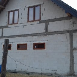 PROFIX-BUDOWNICTWO Krzysztof Jarmułowicz - Surowy stan budynku w trakcie budowy z zamontowanymi brązowymi oknami. Widoczne bloczki, zbrojenia i elementy dachu. W tle budynki mieszkalne i dźwig.