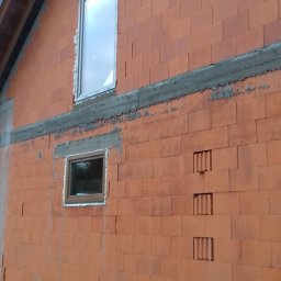 PROFIX-BUDOWNICTWO Krzysztof Jarmułowicz - Montaż okien w surowym budynku z czerwonej cegły. Widoczne dwa okna na różnej wysokości, z pianką montażową. Budynek w trakcie budowy, widoczne niedokończone ściany.