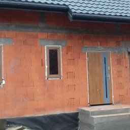 PROFIX-BUDOWNICTWO Krzysztof Jarmułowicz - Montaż stolarki otworowej w surowym budynku: brązowe okna, drzwi zewnętrzne z szarą wstawką i segmentowa brama garażowa w kolorze drewna.