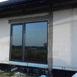 PROFIX-BUDOWNICTWO Krzysztof Jarmułowicz - Ciemne okno w surowym murze z betonu komórkowego, widoczne pianka montażowa i fragment auta. Okno odbija krajobraz. Dom w trakcie budowy, widok z zewnątrz.