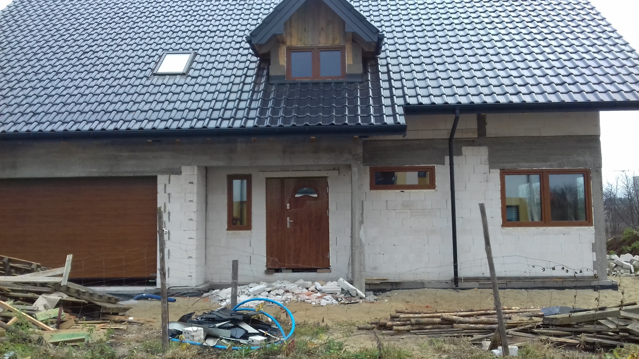 Dom w trakcie budowy w Sulejówku: zamontowane brązowe okna PCV, drzwi wejściowe i brama garażowa. Widoczne materiały budowlane i elementy wykończeniowe.