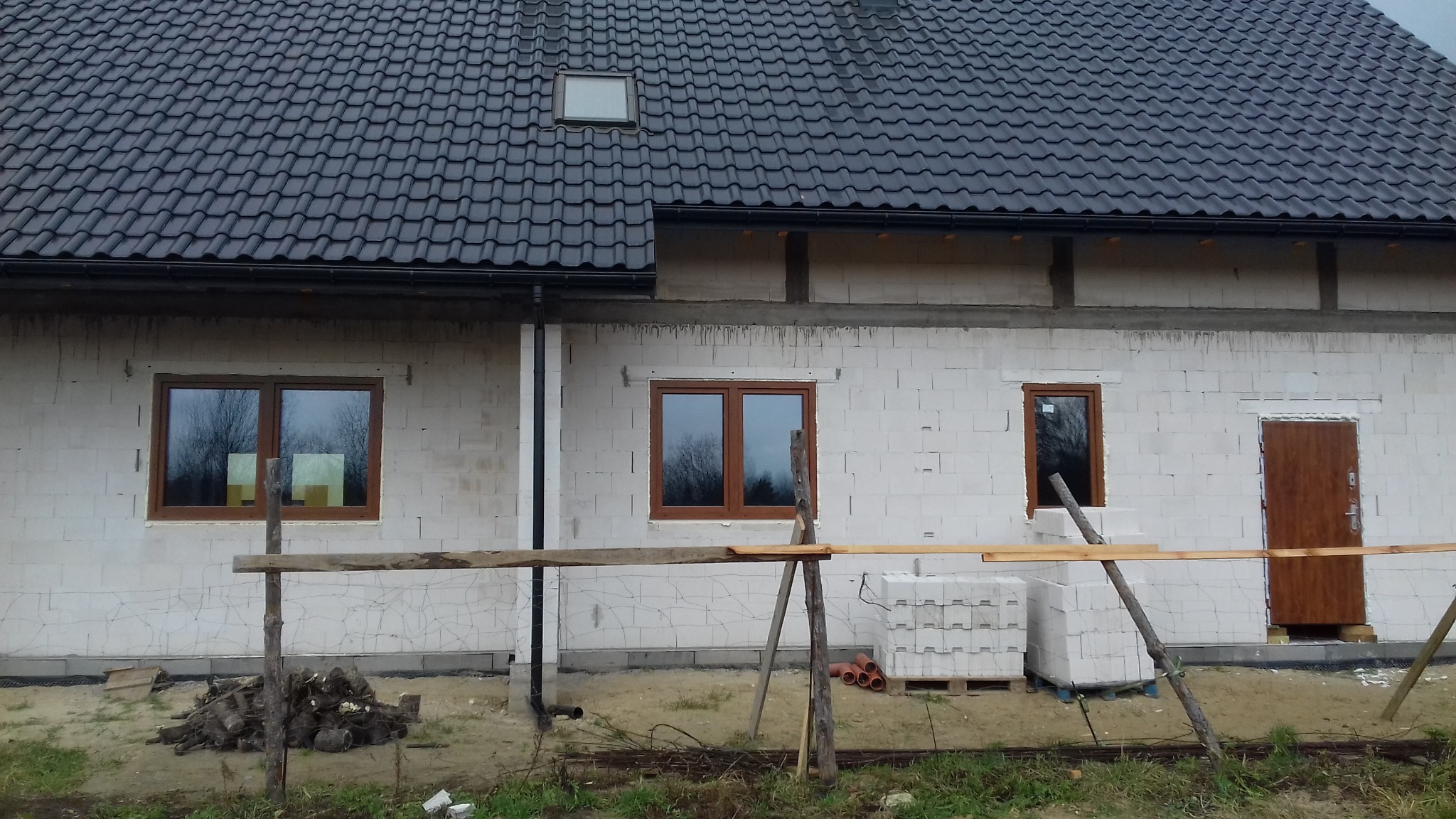 Dom w trakcie budowy z zamontowanymi oknami z brązowymi ramami i drzwiami wejściowymi. Widoczne białe bloczki i czarna dachówka. Częściowo wykonane ocieplenie.