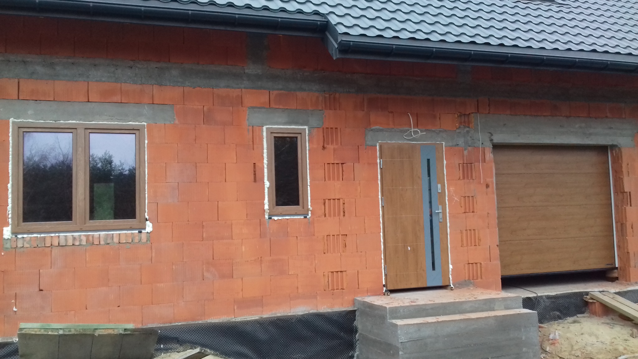 Dom w trakcie budowy: zamontowane okna z brązowymi ramami, drzwi wejściowe z szarą wstawką i brama garażowa w kolorze drewna. Mur z czerwonej cegły.