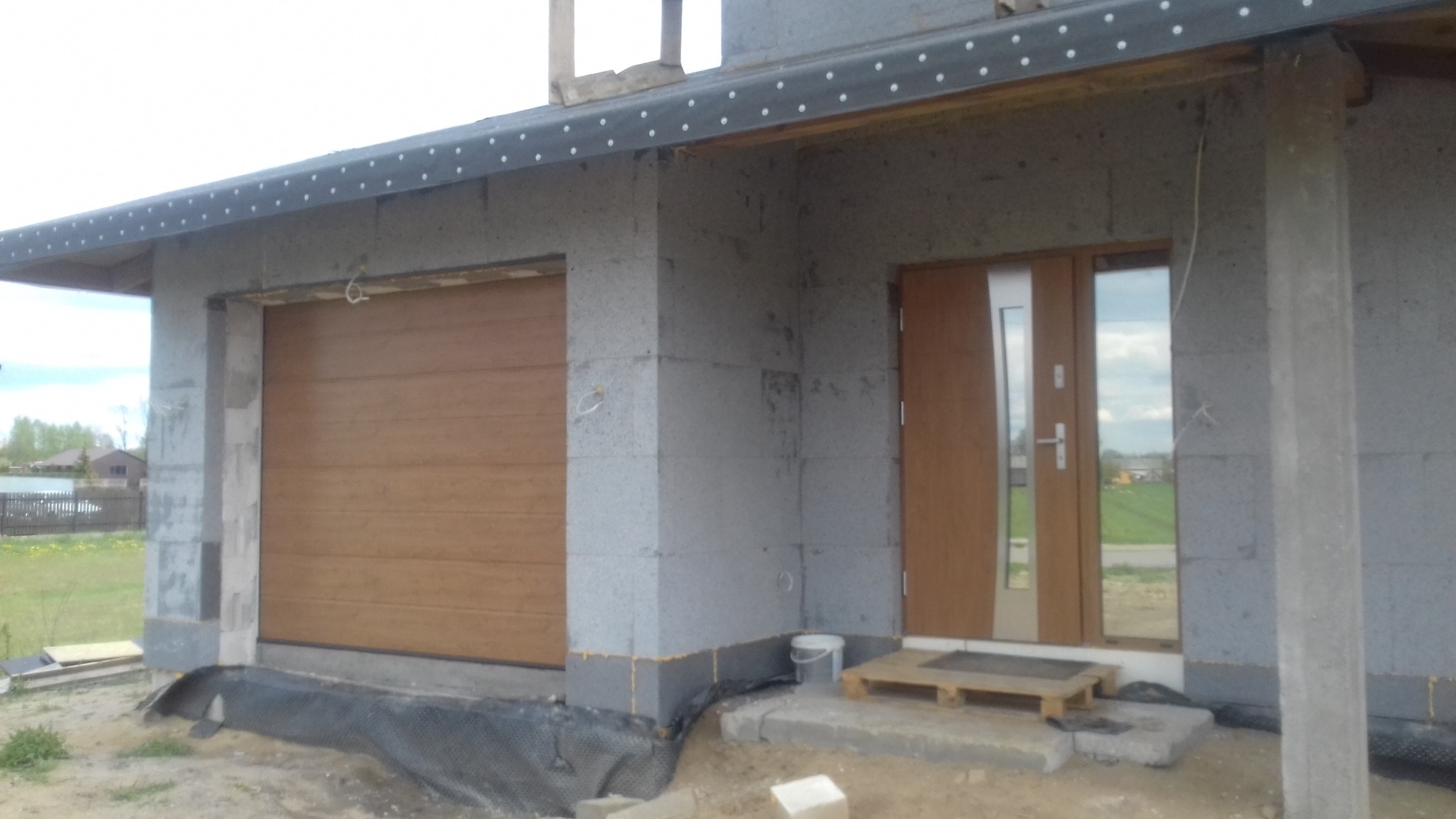 Surowy stan domu w budowie z zamontowaną bramą garażową w kolorze drewna i drzwiami wejściowymi z szybą, widok na okolicę.