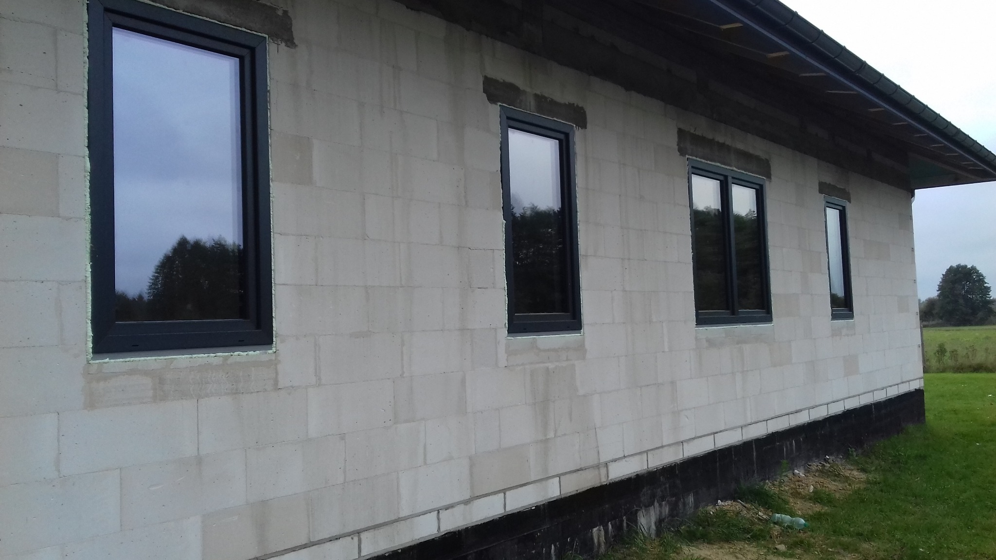 Surowy budynek z szarego betonu komórkowego z zamontowanymi oknami w czarnych ramach. Widok z zewnątrz, w tle zieleń. Etap montażu okien.