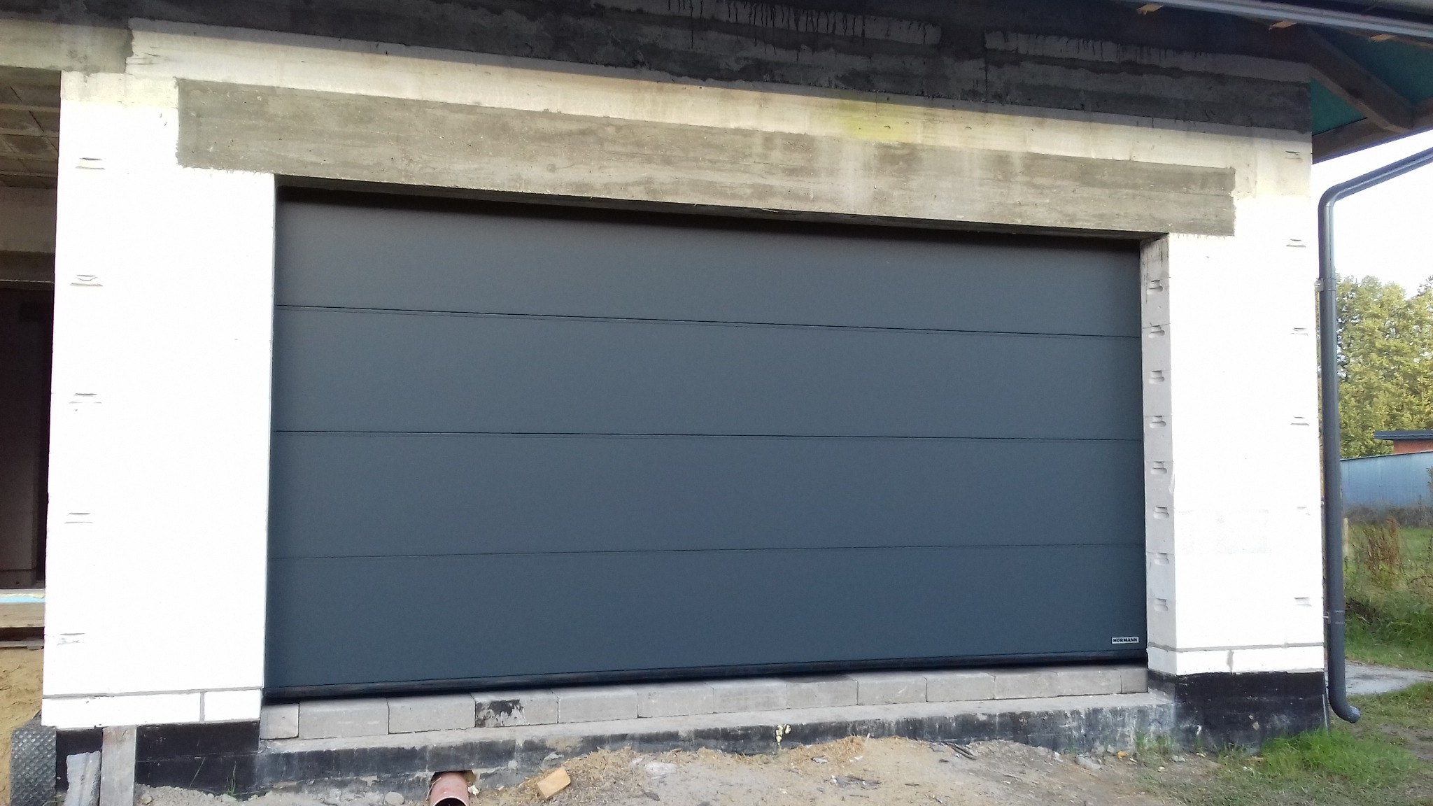 Zamknięta, segmentowa brama garażowa w kolorze grafitowym, zamontowana w surowym otworze budynku. Widoczne białe bloczki i betonowe nadproże.