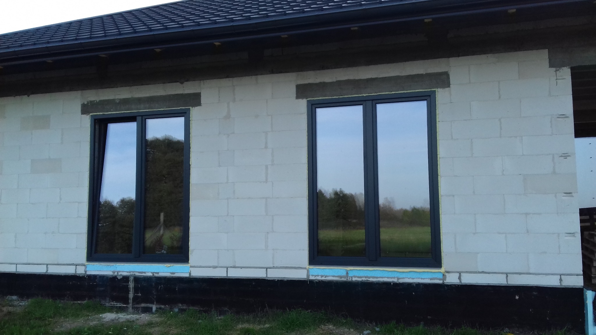 Dwa grafitowe okna w surowym murze z betonu komórkowego. Jedno okno uchylone, widoczne uszczelnienie pianką montażową. W szybach odbija się zieleń drzew.