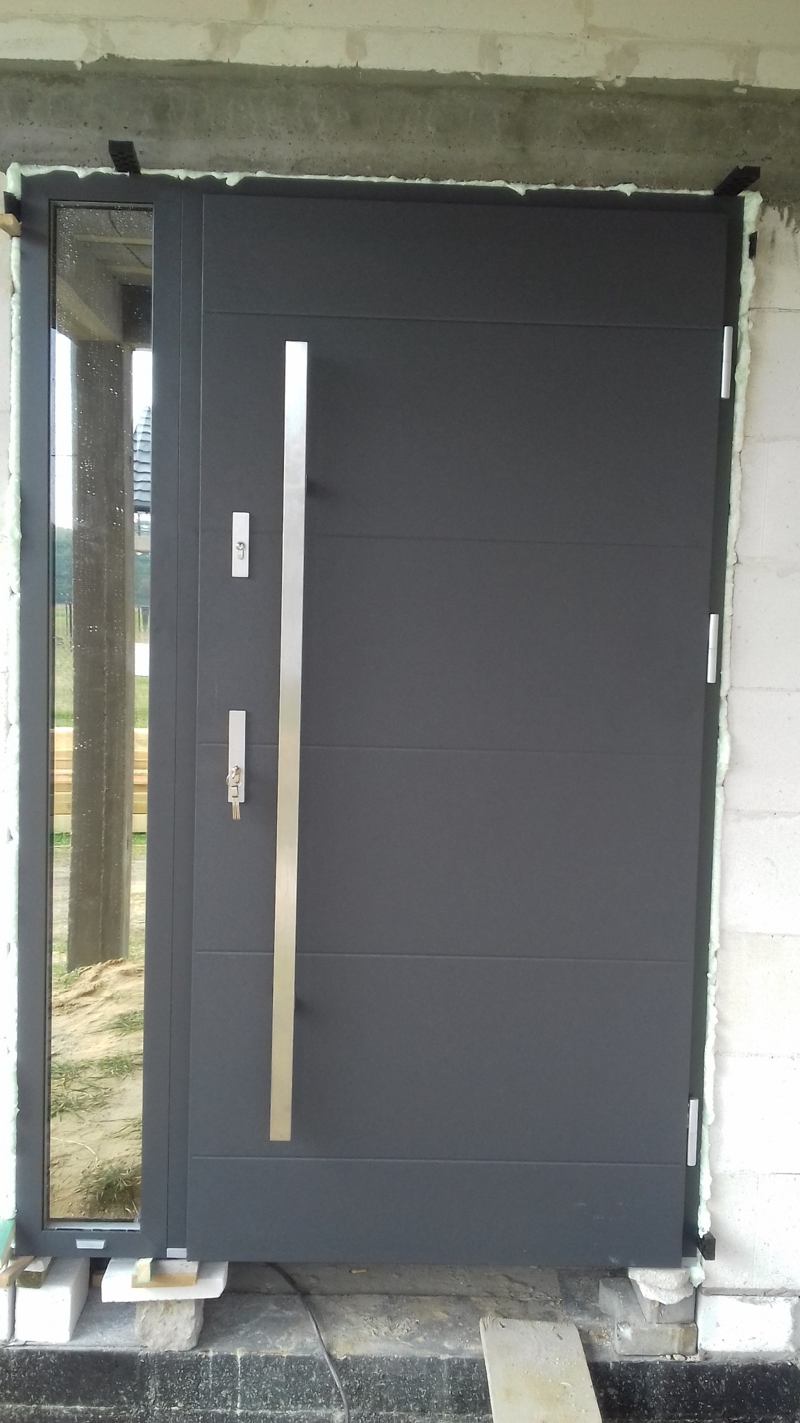 Nowoczesne, grafitowe drzwi zewnętrzne z długim, pionowym uchwytem i szybą boczną, osadzone w surowym otworze, z widoczną pianką montażową i elementami konstrukcyjnymi.