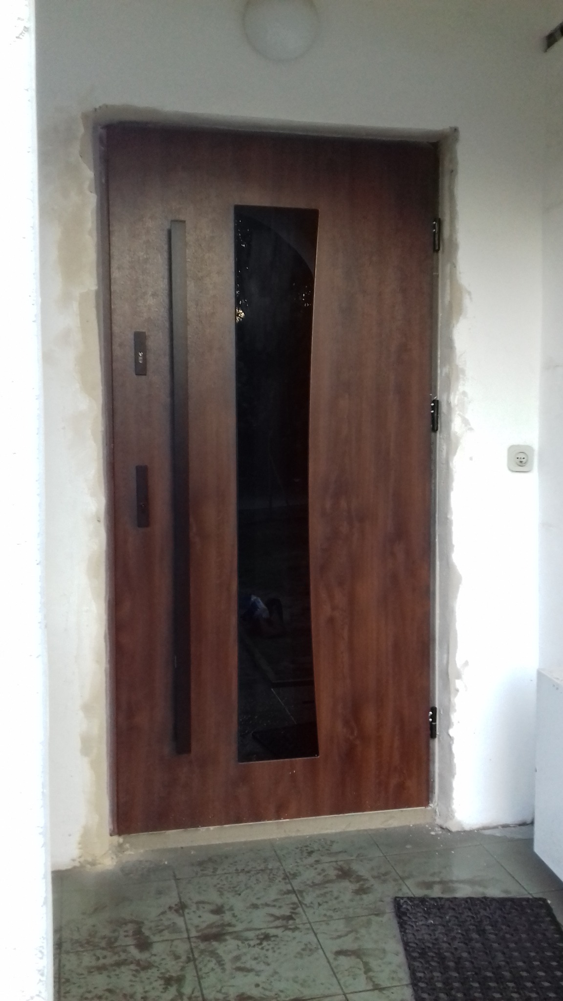 Nowoczesne drzwi wejściowe z drewnopodobnym wykończeniem i pionowym przeszkleniem, osadzone w surowym otworze, czekające na wykończenie ościeżnicy.