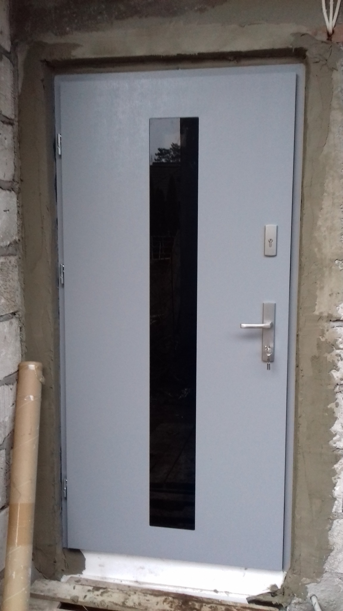 Szare drzwi zewnętrzne z pionowym, prostokątnym przeszkleniem, osadzone w surowym murze. Widoczny zamek, klamka i fragment rury ochronnej po lewej stronie.