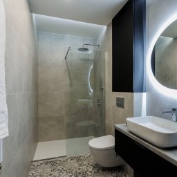 ARVION GROUP SP. Z O.O. - Nowoczesna łazienka z kabiną prysznicową typu walk-in, umywalką nablatową i okrągłym lustrem z podświetleniem LED. Szare płytki i wzorzysta podłoga.