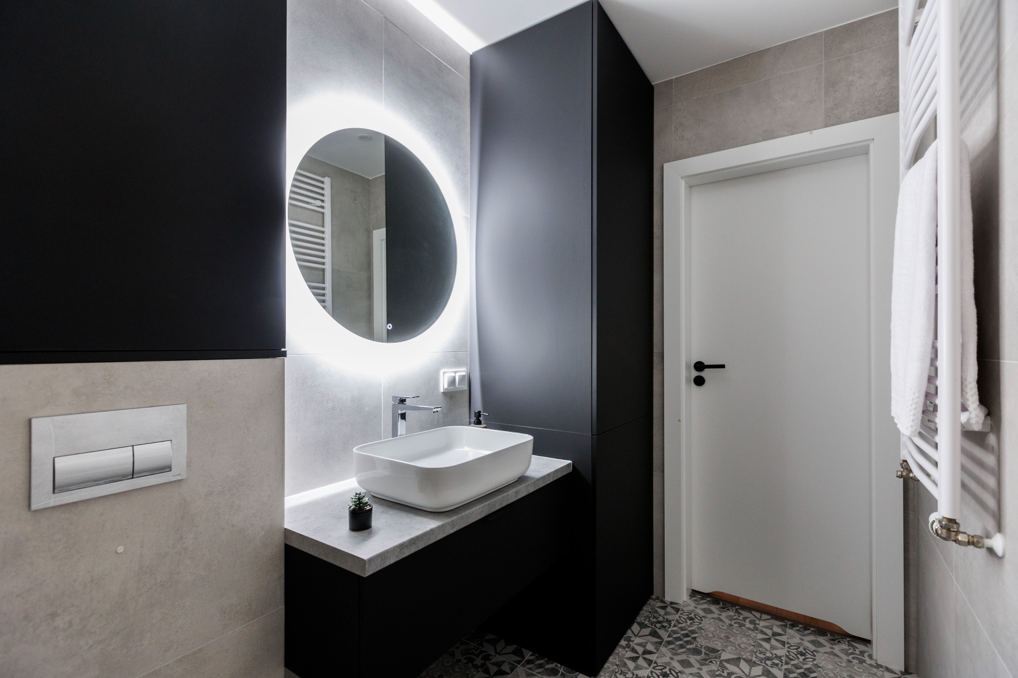 Nowoczesna łazienka z podświetlanym lustrem, białą umywalką na czarnym blacie i wzorzystą podłogą. Minimalistyczny design z szarymi płytkami i białymi akcentami.