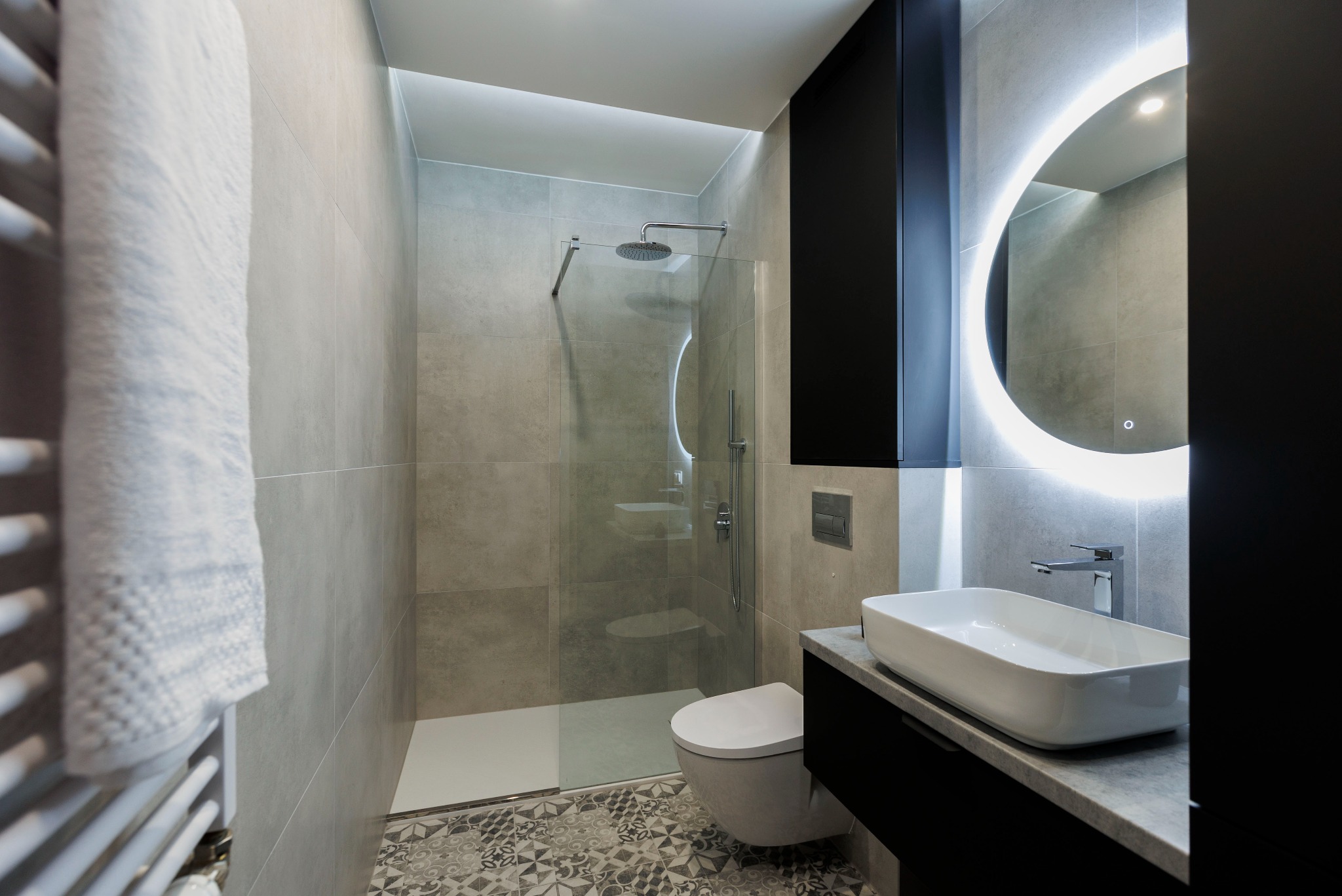 Nowoczesna łazienka z kabiną prysznicową typu walk-in, umywalką nablatową i okrągłym lustrem z podświetleniem LED. Szare płytki i wzorzysta podłoga.