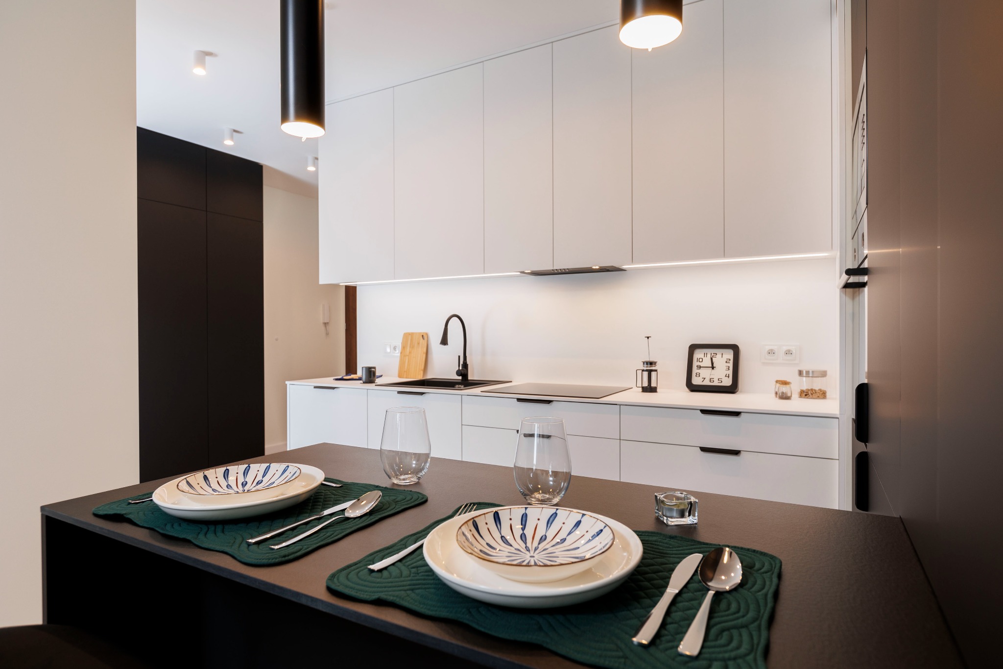 Nowoczesna kuchnia z białymi szafkami i czarnym blatem. Nakryty stół z dekoracyjną zastawą. Minimalistyczne oświetlenie punktowe i wiszące lampy.