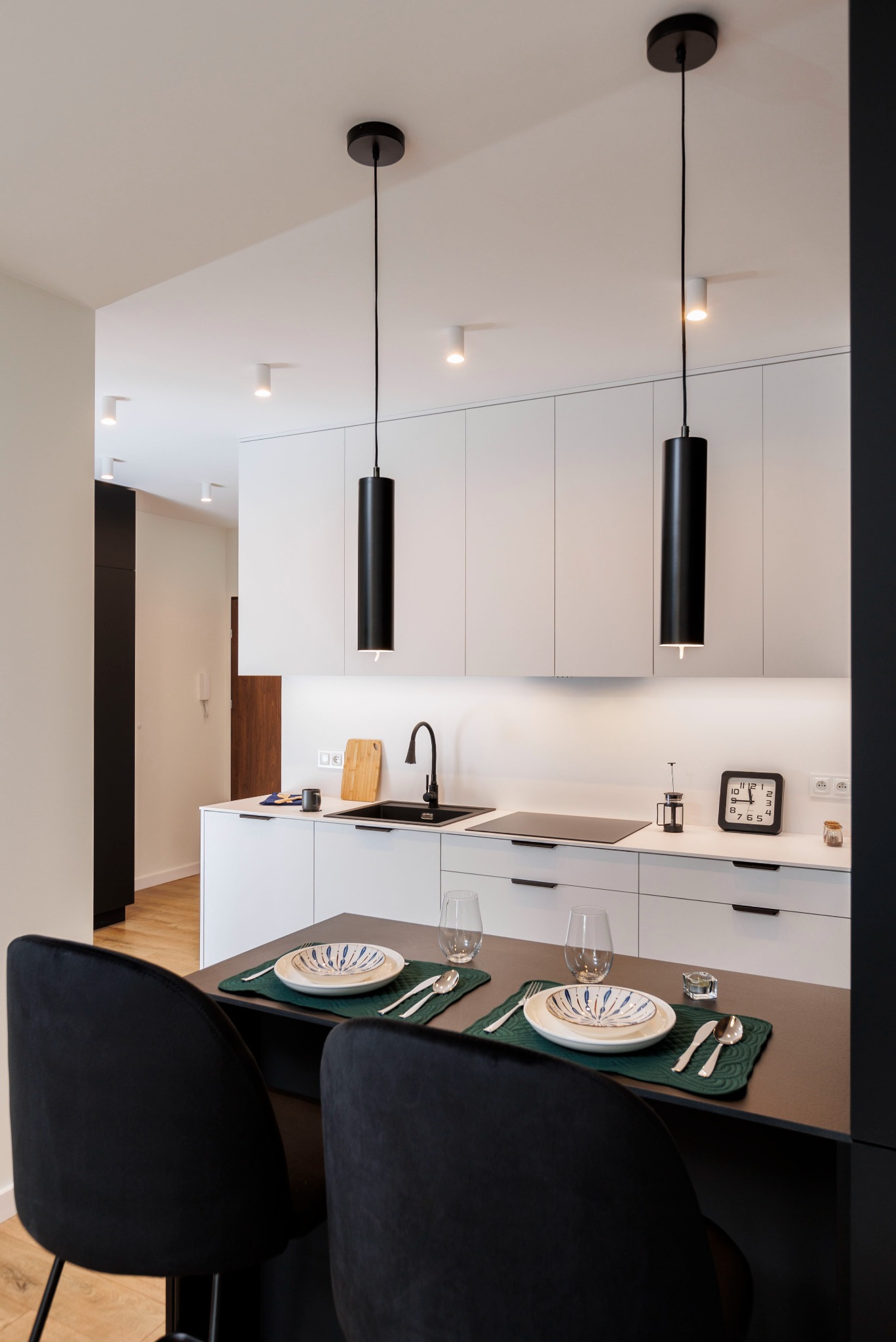 Nowoczesna kuchnia z wyspą i czarnymi lampami cylindrycznymi. Białe szafki, czarne akcenty, nakryty stół. Minimalistyczny design, oświetlenie punktowe.