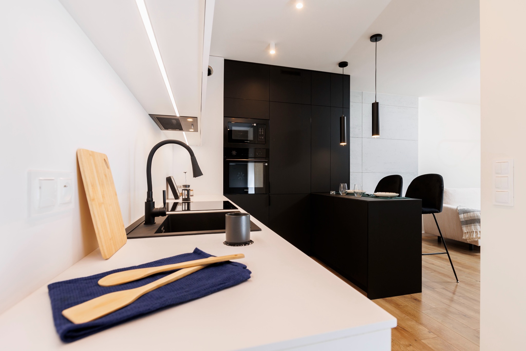 Nowoczesna kuchnia w czerni i bieli z drewnianymi akcentami: deska do krojenia i łyżki na blacie. Minimalistyczny design z oświetleniem punktowym.