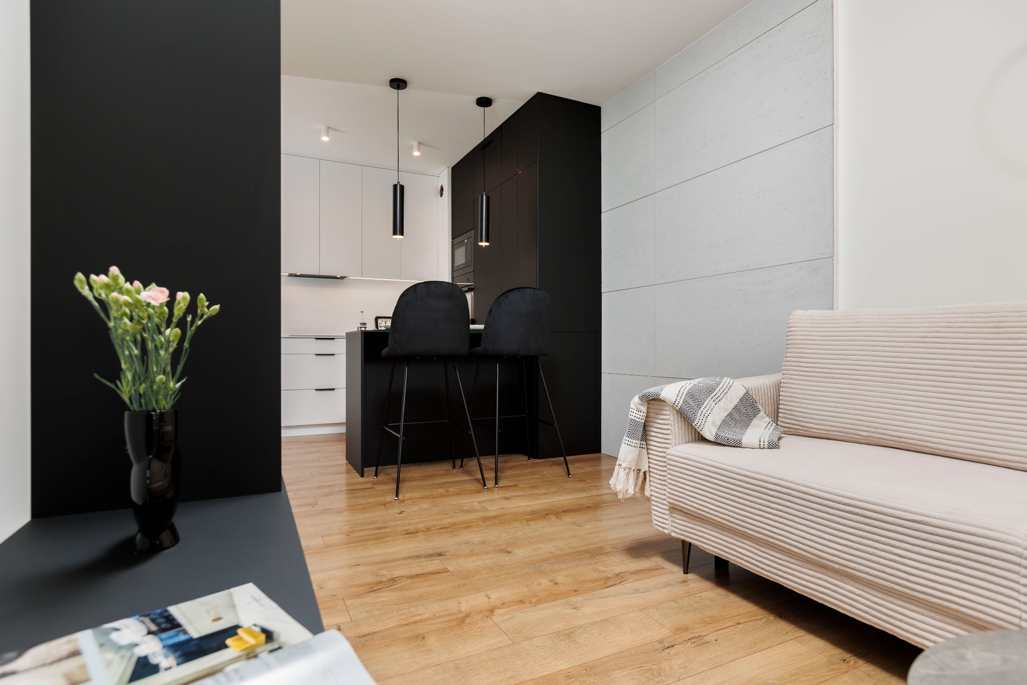 Nowoczesne wnętrze: czarno-biała kuchnia z wyspą i hokerami, drewniana podłoga, beżowa sofa z pledem, kwiaty w wazonie. Minimalistyczny design, eleganckie oświetlenie.