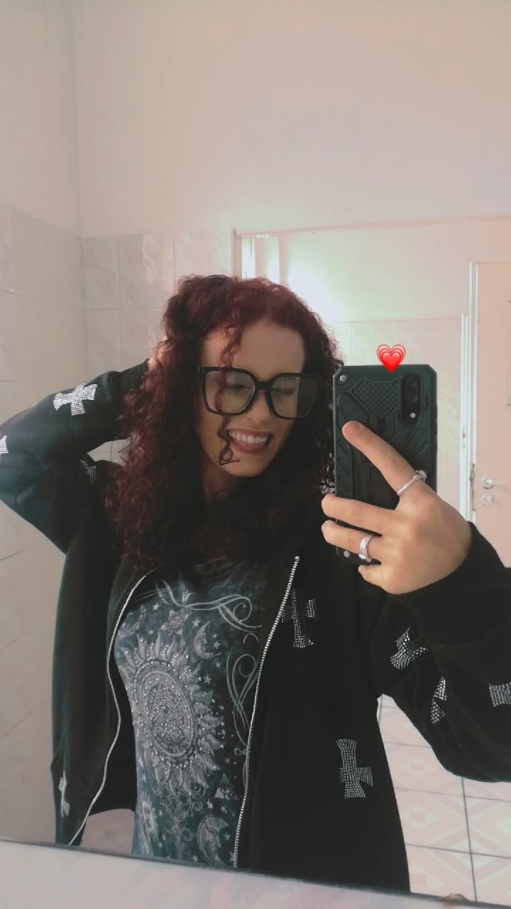 Uśmiechnięta kobieta z kręconymi włosami w okularach robi selfie w łazience, ubrana w bluzę z krzyżami i koszulkę z motywem słońca i księżyca.