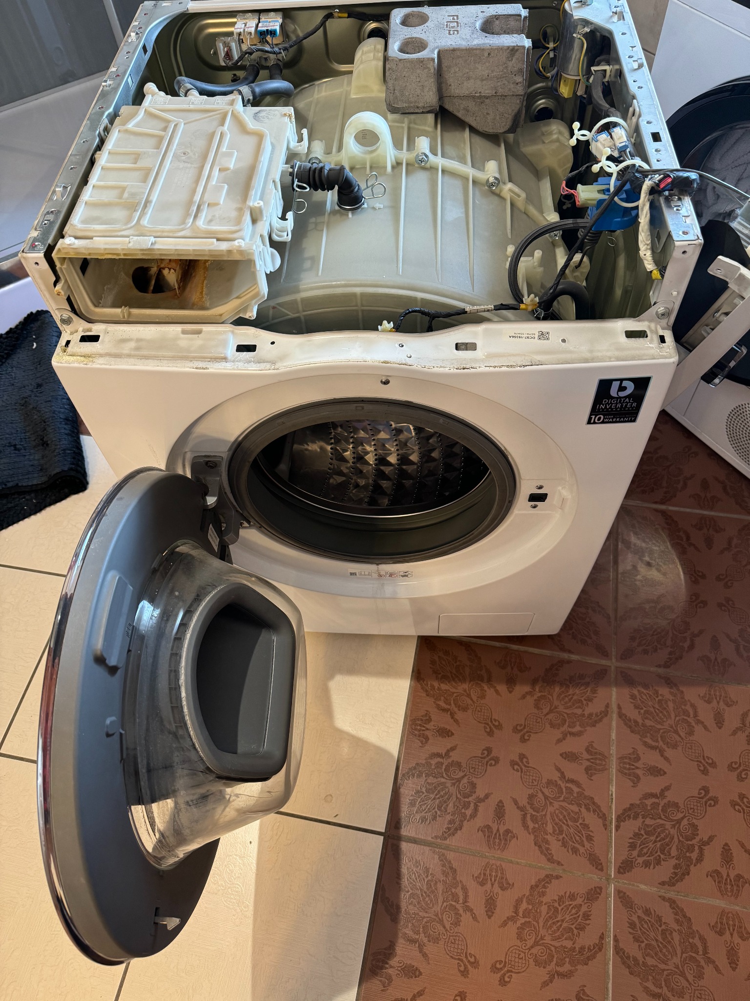 Otwarta pralka z widocznymi elementami wewnętrznymi, w tym bęben i podzespoły. Widoczny otwarty panel przedni. Na podłodze wzorzyste brązowe płytki.