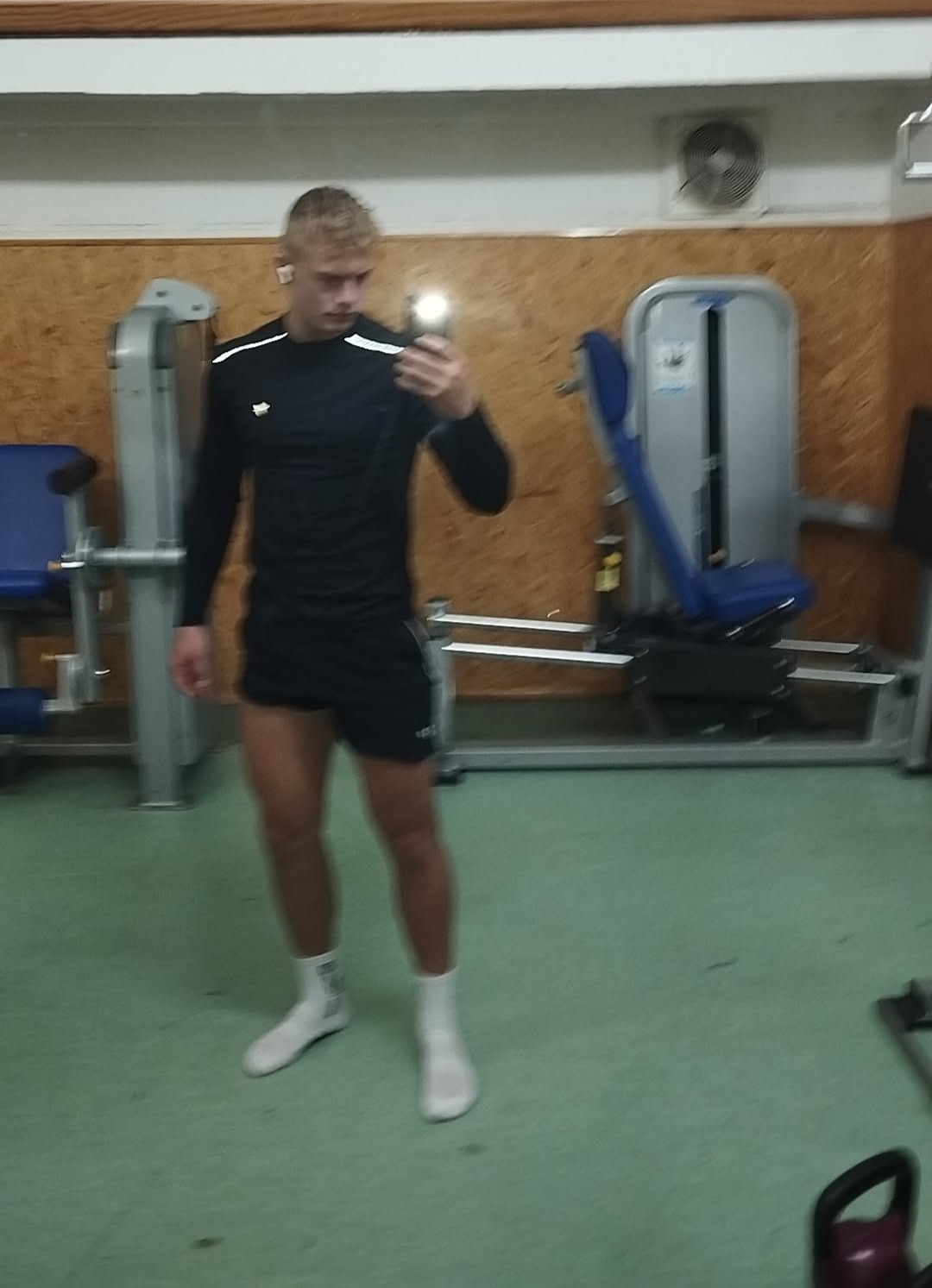 Młody mężczyzna w sportowym stroju robi selfie w siłowni na tle maszyn do ćwiczeń. Ubrany w czarną koszulkę z długim rękawem i krótkie spodenki, białe skarpetki.
