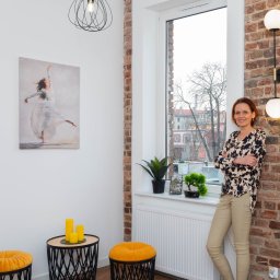 Joanna Kałuska - Kobieta oparta o ceglaną ścianę z lampą w odnowionym wnętrzu. Obraz tancerki, żółte pufy i rośliny doniczkowe dodają charakteru. Widok z okna na miasto.