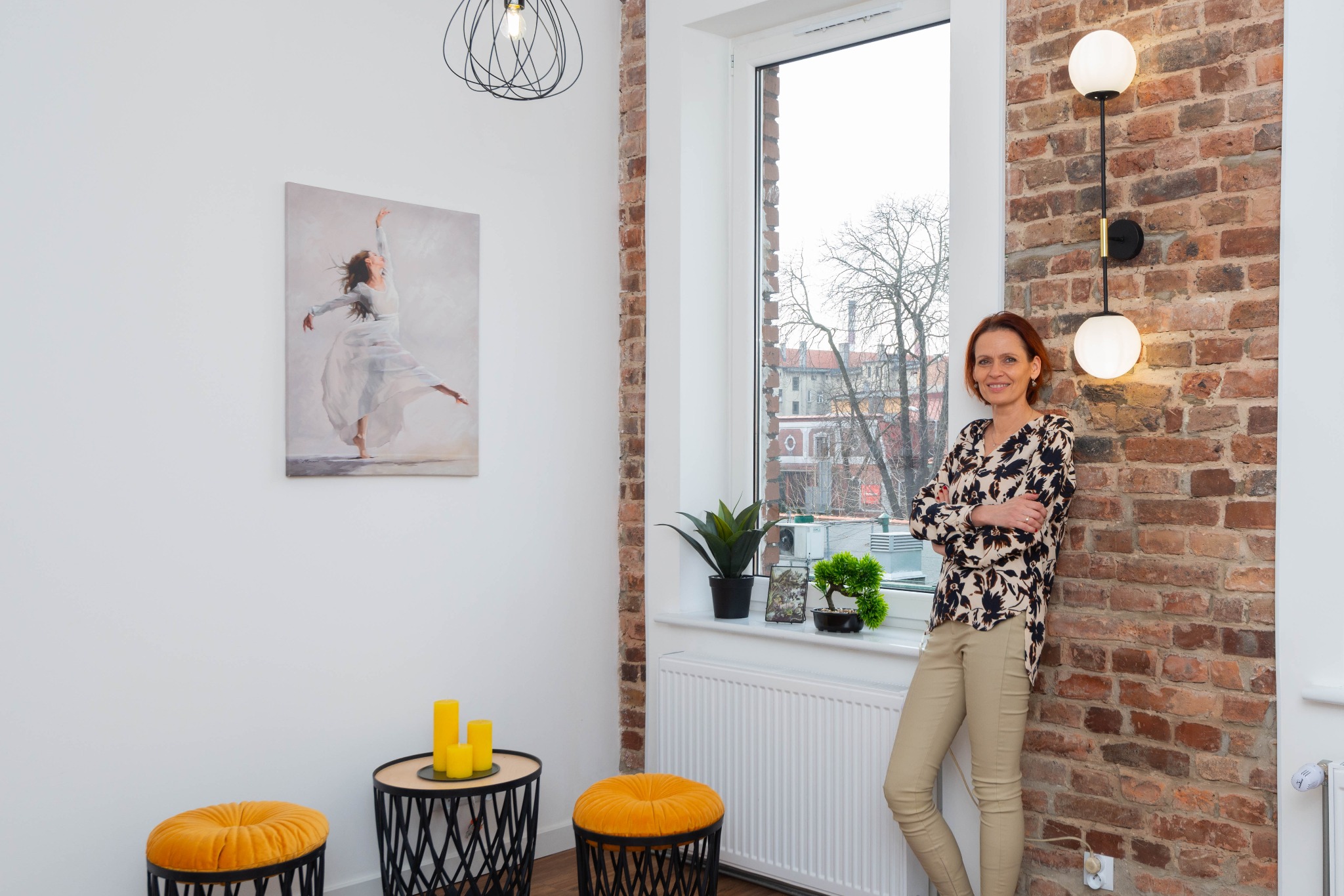 Kobieta oparta o ceglaną ścianę z lampą w odnowionym wnętrzu. Obraz tancerki, żółte pufy i rośliny doniczkowe dodają charakteru. Widok z okna na miasto.