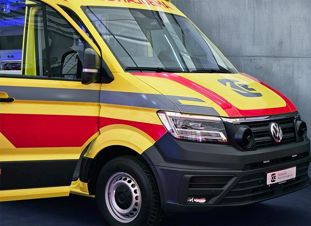 Żółto-czerwony ambulans Volkswagen Crafter z widocznym logo Special Performance na tle betonowej ściany. Ujęcie z przodu, skupione na detalu.