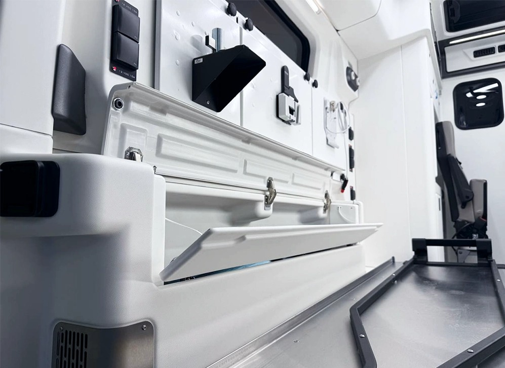 Wnętrze specjalistycznego pojazdu z otwartą, białą szafką i elementami wyposażenia, w tym czarne przełączniki i uchwyty, sugerujące zaawansowaną instalację elektryczną.