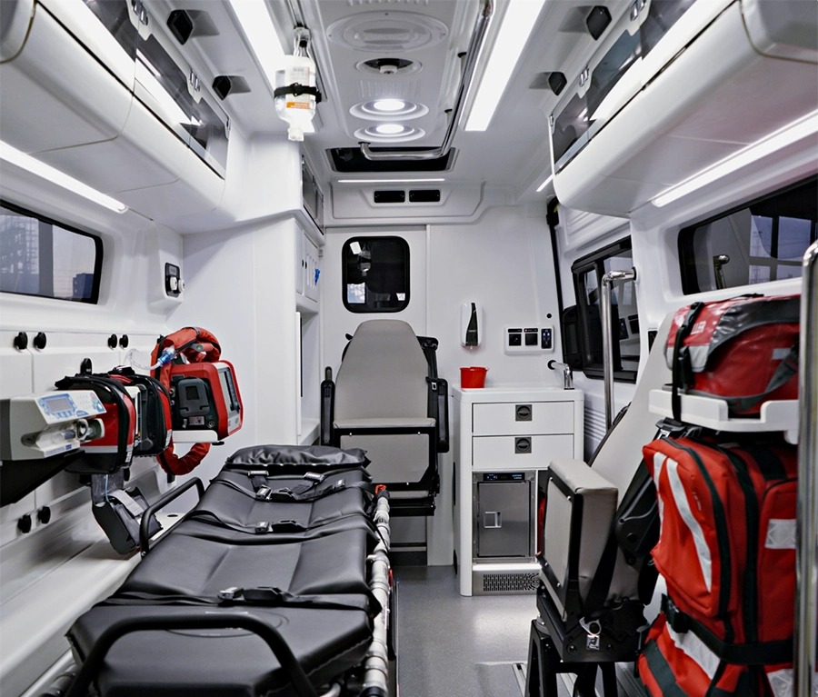 Wnętrze ambulansu z noszami, torbami medycznymi i sprzętem. Widok z tyłu pojazdu, oświetlenie sufitowe, białe ściany i wyposażenie.