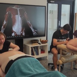 HEROS CENTRUM SZKOLENIOWE Adam Rudowski - Kurs masażu w Warszawie: Uczestnicy szkolenia praktykują techniki na modelach, w tle ekran z anatomią, szkielet i apteczka pierwszej pomocy.