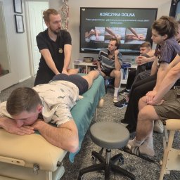 HEROS CENTRUM SZKOLENIOWE Adam Rudowski - Kurs masażu: instruktor demonstruje techniki na leżącym uczestniku, grupa obserwuje. Ekran z ilustracjami, szkielet anatomiczny w tle. Jasne wnętrze, profesjonalny sprzęt.