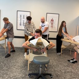 HEROS CENTRUM SZKOLENIOWE Adam Rudowski - Kurs masażu: grupa osób ćwiczy techniki masażu na leżankach w jasnym pomieszczeniu. Na ścianach wiszą plakaty z anatomią człowieka. Widoczny fotel na kółkach.