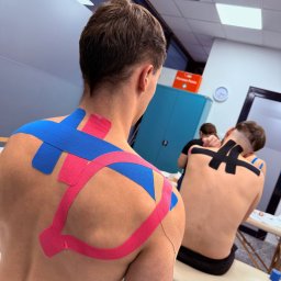 HEROS CENTRUM SZKOLENIOWE Adam Rudowski - Kinesiotaping pleców dwóch mężczyzn w sali szkoleniowej. Kolorowe taśmy (różowe, niebieskie, czarne) aplikowane na skórę. Widoczny stół do masażu i szafka.