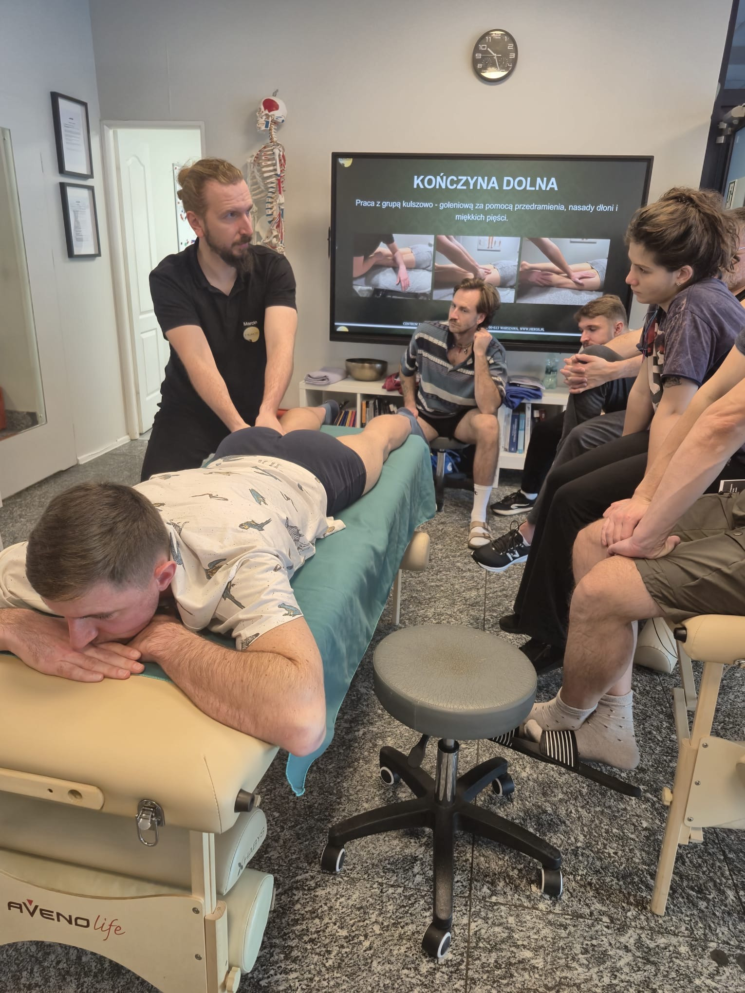 Kurs masażu: instruktor demonstruje techniki na leżącym uczestniku, grupa obserwuje. Ekran z ilustracjami, szkielet anatomiczny w tle. Jasne wnętrze, profesjonalny sprzęt.