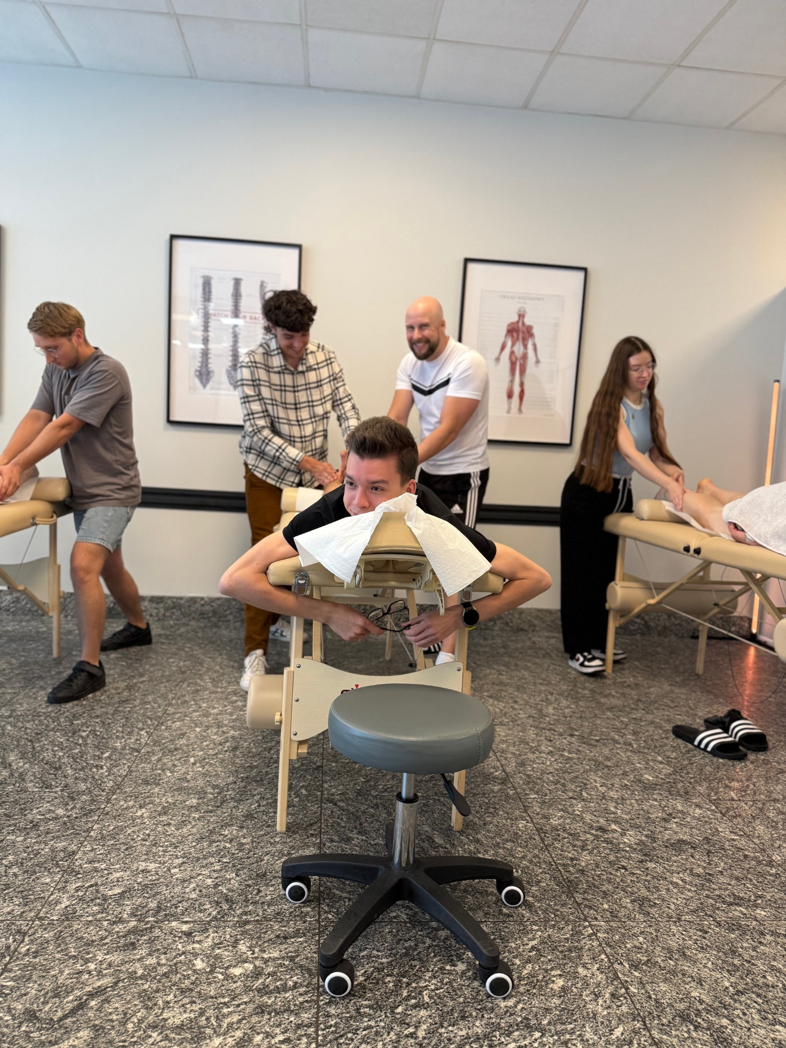 Kurs masażu: grupa osób ćwiczy techniki masażu na leżankach w jasnym pomieszczeniu. Na ścianach wiszą plakaty z anatomią człowieka. Widoczny fotel na kółkach.