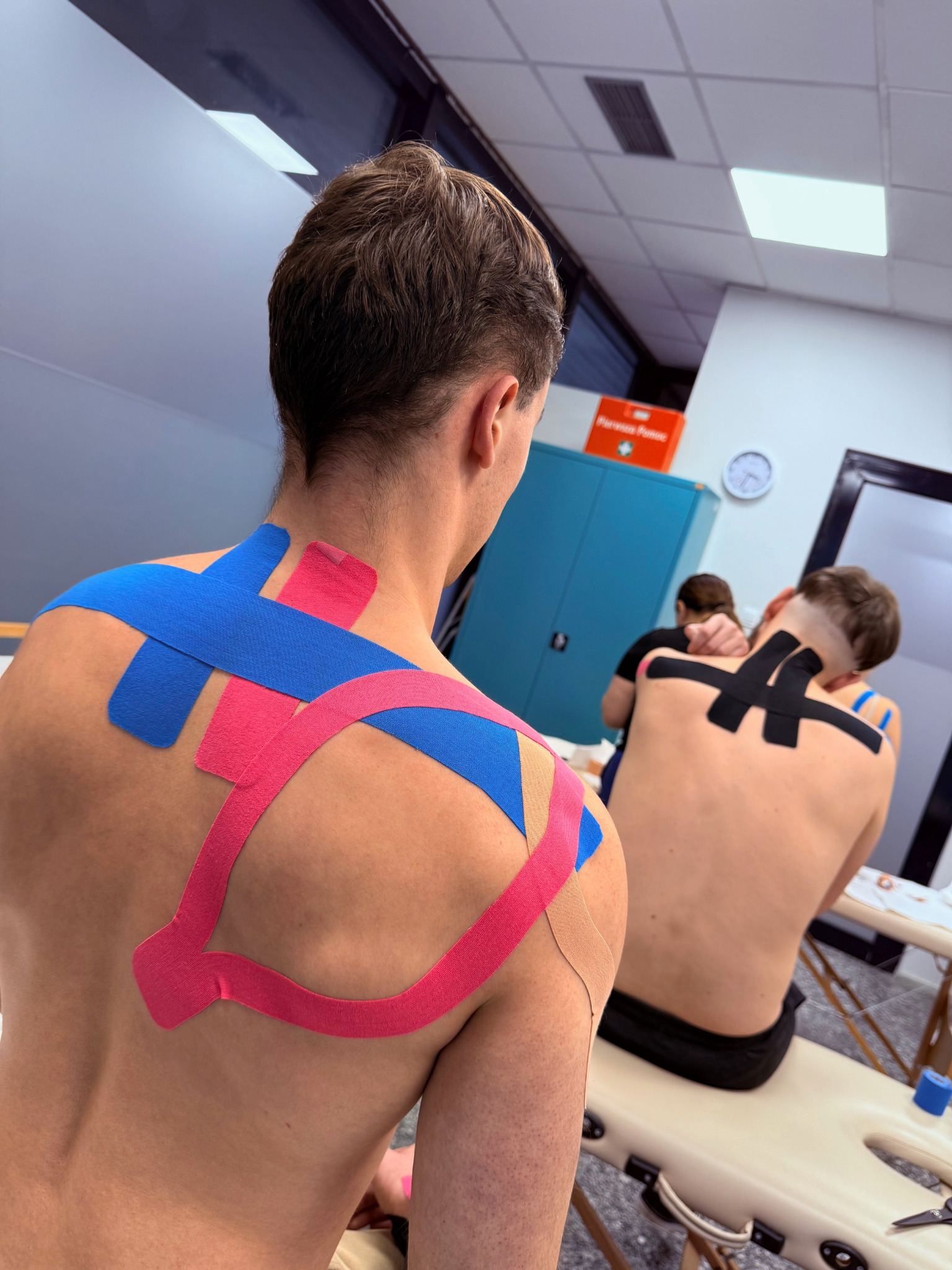 Kinesiotaping pleców dwóch mężczyzn w sali szkoleniowej. Kolorowe taśmy (różowe, niebieskie, czarne) aplikowane na skórę. Widoczny stół do masażu i szafka.
