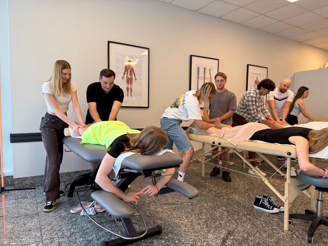 Sala szkoleniowa: kursanci pod okiem instruktorów ćwiczą masaż na leżących modelach. Widoczne plakaty anatomiczne na ścianie i profesjonalne stoły do masażu.