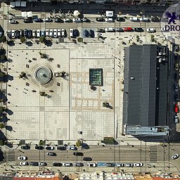 DRONAM dron*foto*film - Widok z drona na rynek miejski z fontanną, budynkami i zaparkowanymi samochodami. Geometryczny układ placu, cień rzucany przez budynek. Logo firmy w prawym górnym rogu.