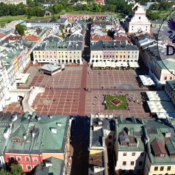 DRONAM dron*foto*film - Panorama Zamościa z lotu ptaka, ukazująca Rynek Wielki z kolorowymi kamienicami, ratuszem i katedrą. Widoczny logotyp firmy Dronam.