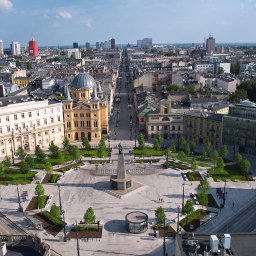 DRONAM dron*foto*film - Panorama miasta z lotu ptaka, centralny plac z fontanną i budynkami w stylu klasycystycznym, w tle nowoczesne wieżowce. Logo firmy w prawym górnym rogu.
