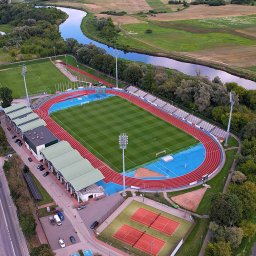 DRONAM dron*foto*film - Widok z drona na stadion lekkoatletyczny z bieżnią, boiskiem, trybunami i kortami tenisowymi, położony nad rzeką, otoczony zielenią. W tle droga i zabudowania.